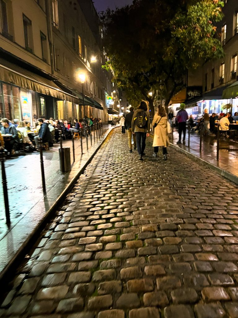Evening in a Paris passage. Photo has people dining, others walking on the cobblestone path.
