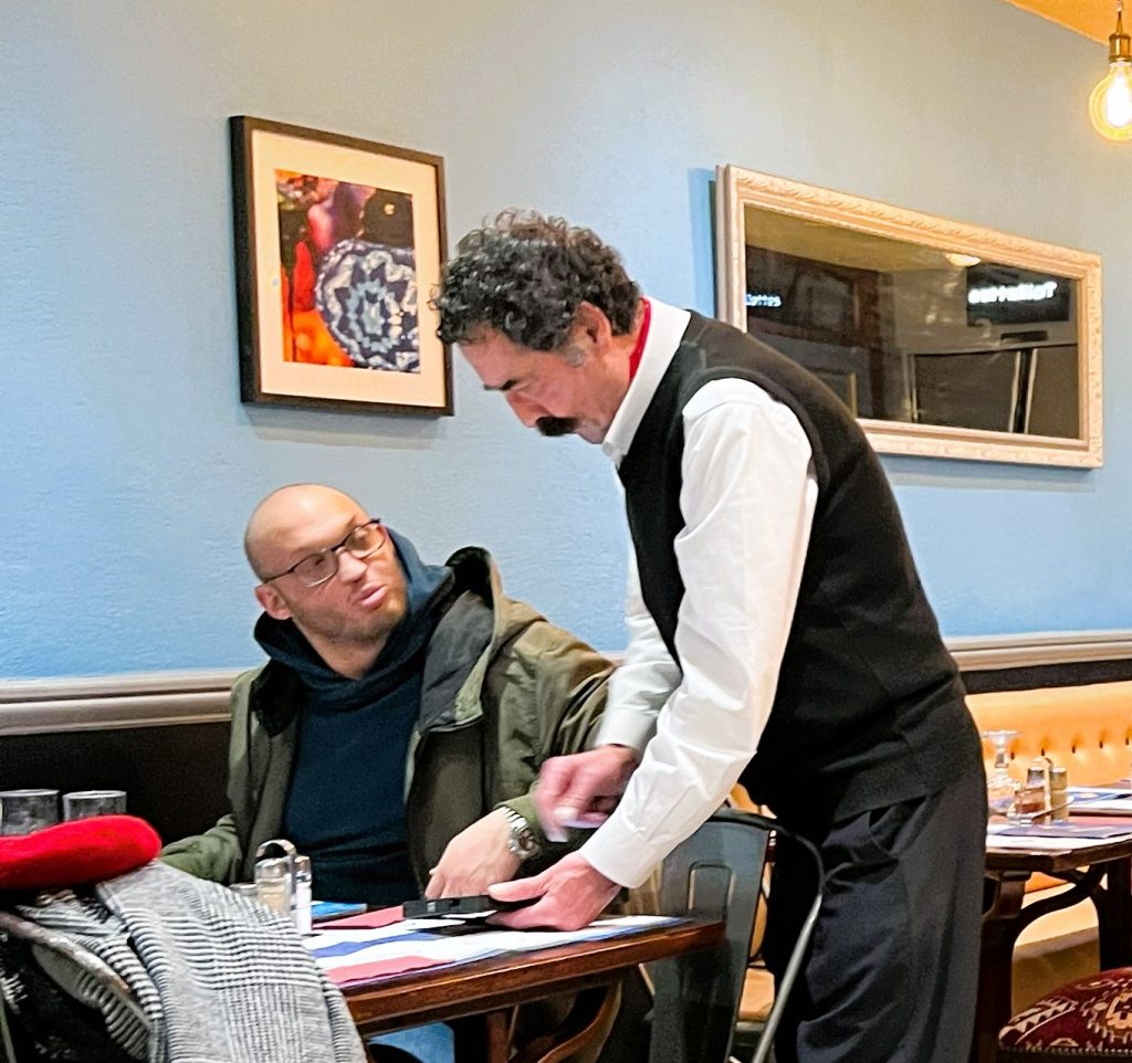 a photo of a waiter with a very large moustache taking the order of another gentleman with glasses on and a coat and red beret laying on the table